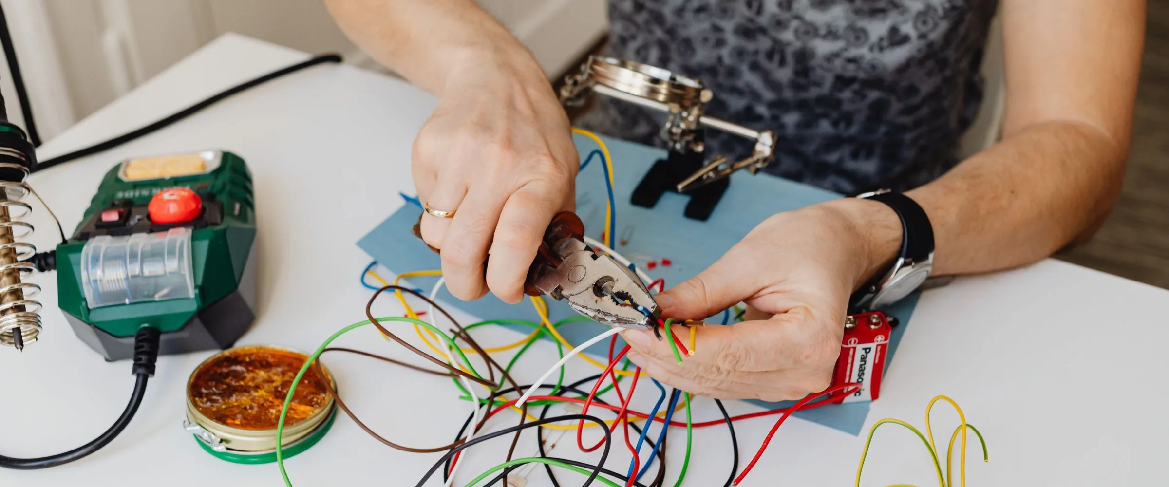 Hands stripping electrical wire at a workbench, beside a soldering station
